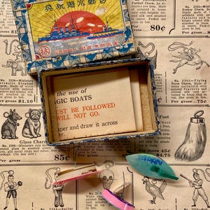 May include: Vintage box with a colorful illustration of a ship and text in a foreign language. Inside, a card reads "the use of magic boats must be followed will not go." Several small charms are scattered around.