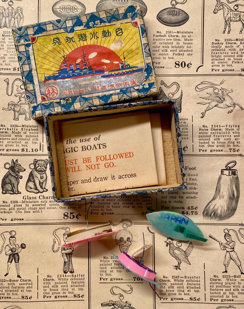 May include: Vintage box with a colorful illustration of a ship and text in a foreign language. Inside, a card reads "the use of magic boats must be followed will not go." Several small charms are scattered around.