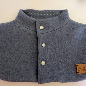 May include: A blue-grey wool collar with a high, rounded neckline and three white buttons down the front. The inside collar lining features a white polka dot pattern on a dark grey background. A small, square, brown leather tag is sewn on the lower right.