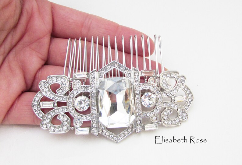 May include: Silver hair comb with a large clear crystal and smaller crystals around the edges. The comb has a decorative design with a repeating pattern.