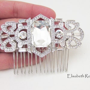 May include: Silver hair comb with a large clear crystal in the center and smaller crystals around the edges. The comb has a decorative design with a geometric pattern.