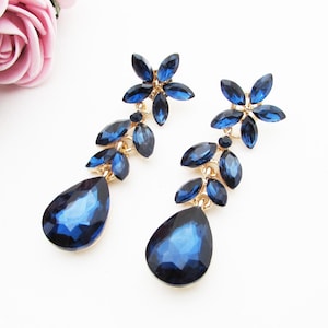Puede incluir: Un par de pendientes largos y colgantes con un diseño de flor de cristal azul. Los pendientes presentan un cristal en forma de lágrima en la parte inferior y cristales más pequeños dispuestos en forma de flor en la parte superior.