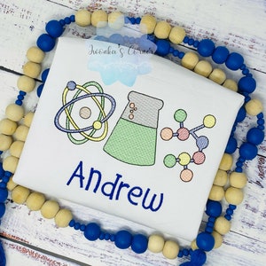 Könnte beinhalten: Weißes T-Shirt mit einem blauen gestickten Namen "Andrew" und bunten, wissenschaftlich inspirierten Illustrationen, darunter ein Atom, ein Becherglas und ein Molekül.