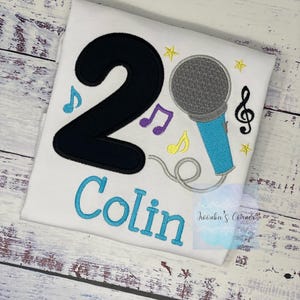 May include: White t-shirt with a black number 2, a blue microphone, and the name "Colin" in blue embroidery. Musical notes and stars in yellow, purple, and blue decorate the shirt, which is likely for a second birthday.