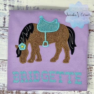 Puede incluir: Una camiseta morada con un caballo marr&oacute;n con una silla de montar turquesa y una flor amarilla en la cabeza. El nombre "BRIDGETTE" est&aacute; bordado en turquesa debajo del caballo.