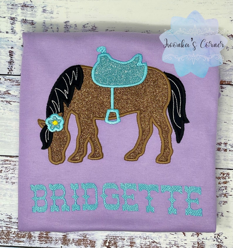 Puede incluir: Una camiseta morada con un caballo marr&oacute;n con una silla de montar turquesa y una flor amarilla en la cabeza. El nombre "BRIDGETTE" est&aacute; bordado en turquesa debajo del caballo.