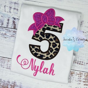 K&ouml;nnte beinhalten: Wei&szlig;es T-Shirt mit einer rosa Schleife und einer Leopardenmuster-Nummer 5. Der Name "Nylah" ist in rosa unter der Nummer gestickt.