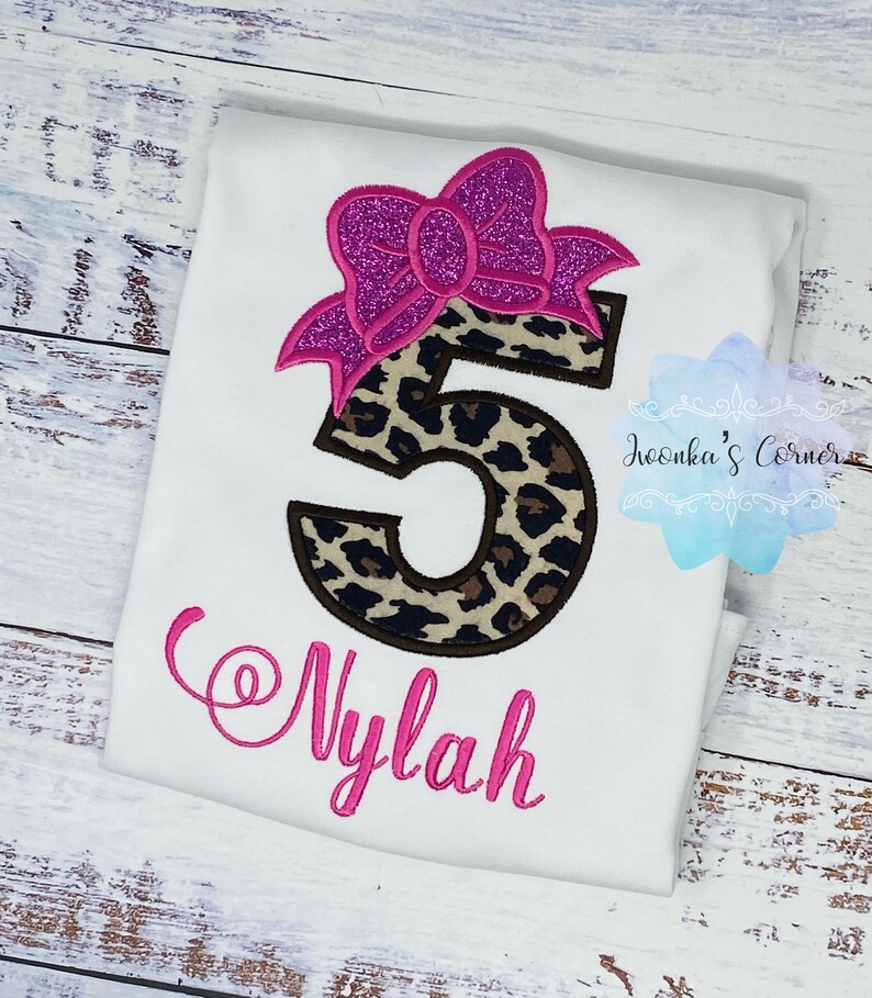 K&ouml;nnte beinhalten: Wei&szlig;es T-Shirt mit einer rosa Schleife und einer Leopardenmuster-Nummer 5. Der Name "Nylah" ist in rosa unter der Nummer gestickt.