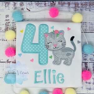 Op de afbeelding: Wit T-shirt met een turquoise nummer 4 met witte stippen en een grijze kat met een bloemenkrans. De naam "Ellie" is turquoise geborduurd. Roze, gele en blauwe pompons zijn rond het shirt geregen.