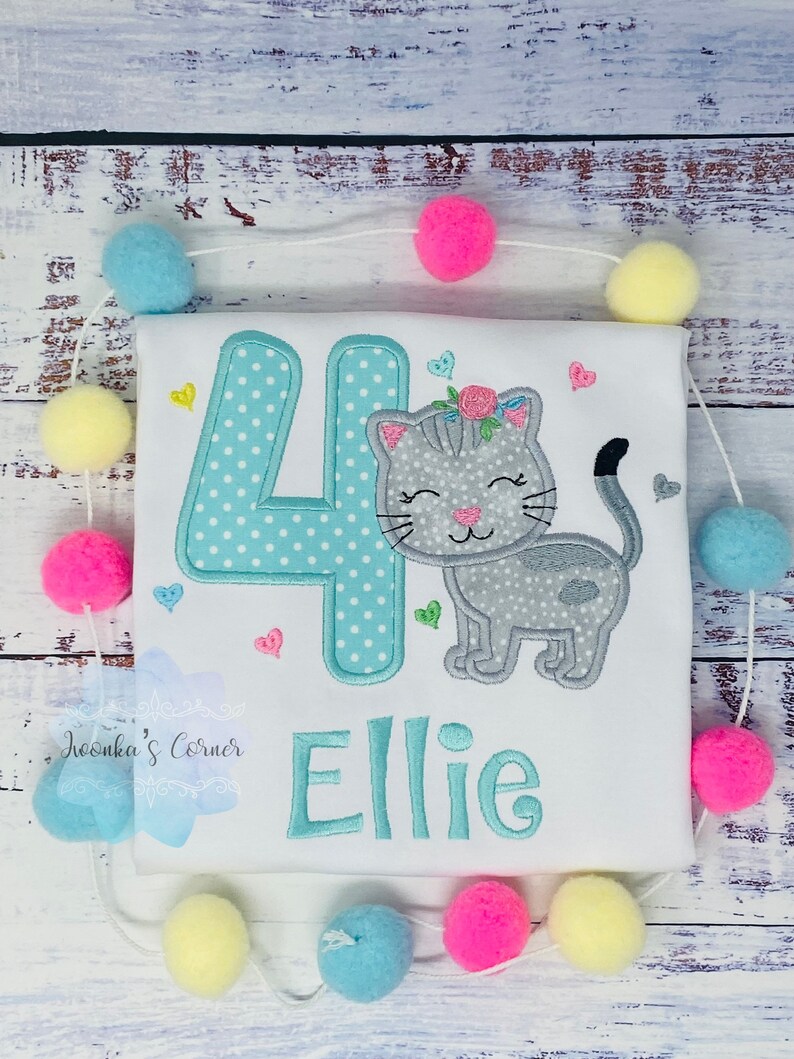 Op de afbeelding: Wit T-shirt met een turquoise nummer 4 met witte stippen en een grijze kat met een bloemenkrans. De naam "Ellie" is turquoise geborduurd. Roze, gele en blauwe pompons zijn rond het shirt geregen.