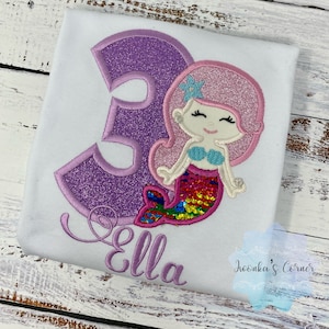 May include: Purple glittery number three with a mermaid design. The mermaid has pink hair, a blue seashell bra, and a colorful sequin tail. The name "Ella" is embroidered below the number.
