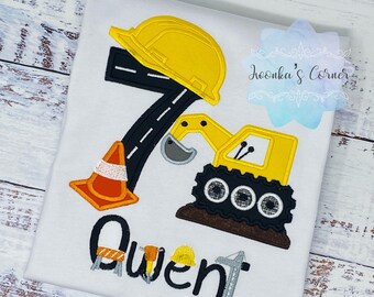 May include: A white t-shirt with an embroidered design of a yellow construction vehicle, a traffic cone, a hard hat, and the number 7. The name "Owen" is embroidered below the design.