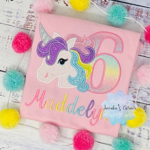 May include: Pink t-shirt with a rainbow glitter unicorn and the number 6. The name "Maddelyn" is embroidered in rainbow colors below the unicorn. The text "Wonka's Corner" is in the bottom right corner.