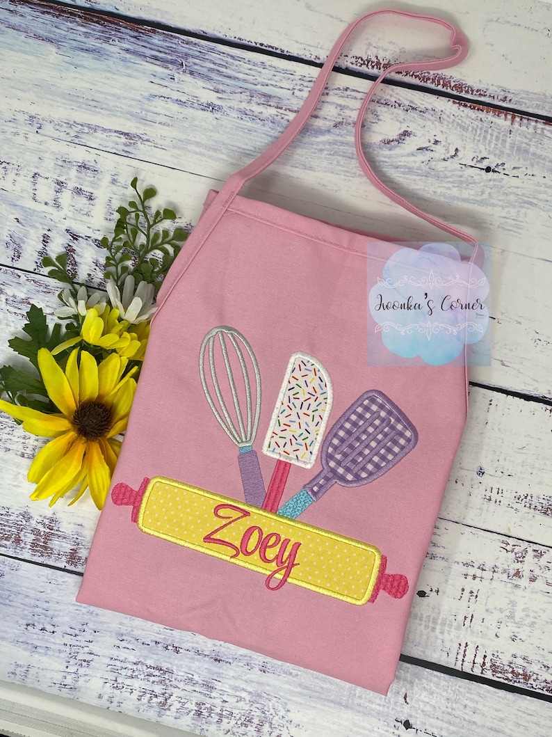 May include: Pink child's apron with a yellow rolling pin, a whisk, a spatula, and sprinkles. The rolling pin has the name "Zoey" embroidered on it.