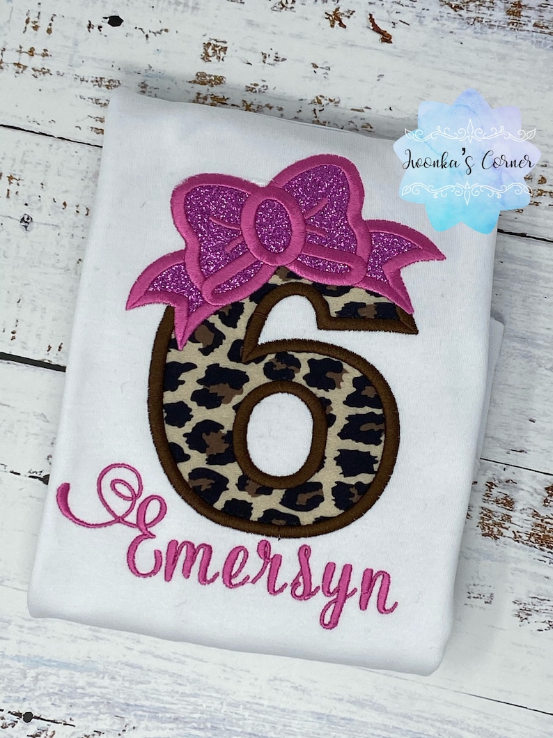 K&ouml;nnte beinhalten: Ein wei&szlig;es Shirt mit einer rosa Schleife und einer braunen und schwarzen Leopardenmuster-Zahl sechs. Der Name "Emersyn" ist in rosa unter der Zahl gestickt.