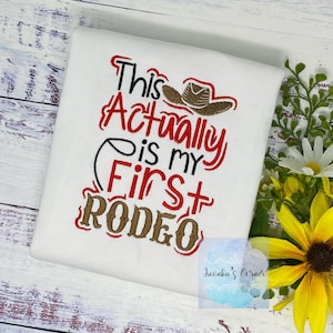 Könnte beinhalten: Weißes T-Shirt mit roter und brauner Stickerei, auf dem "This Actually is my First Rodeo" steht, mit einem braunen Cowboyhut über dem Text.