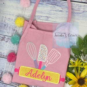 May include: Pink child's apron with a yellow rolling pin and three kitchen tools embroidered on it. The name "Adelyn" is embroidered on the rolling pin.