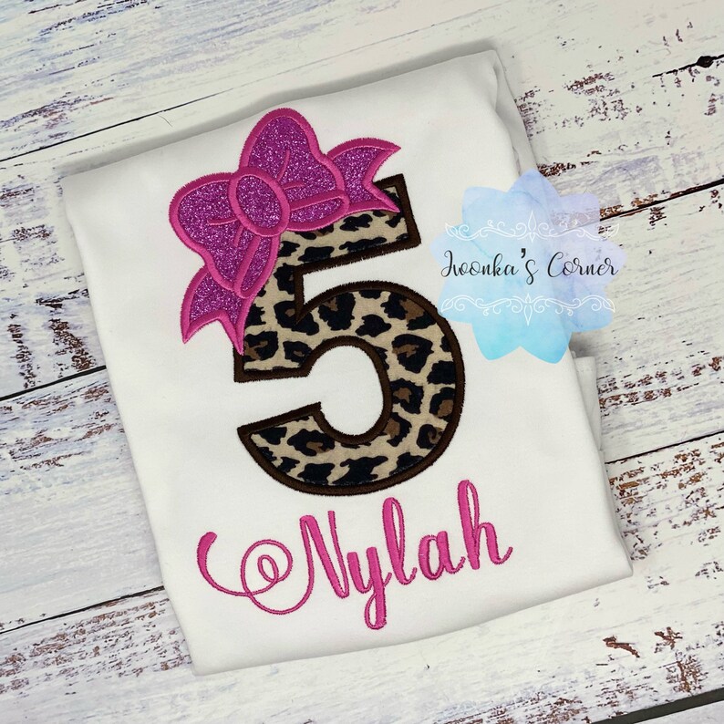 K&ouml;nnte beinhalten: Ein wei&szlig;es T-Shirt mit einer rosa Schleife und einer Leopardenmuster-Nummer 5. Der Name "Nylah" ist in rosa unter der Nummer gestickt.
