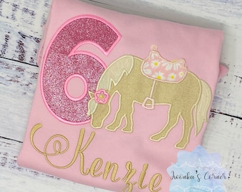 May include: Pink shirt with a glittery pink number six, a gold embroidered horse with a saddle, and the name "Kenzie" in gold embroidery.