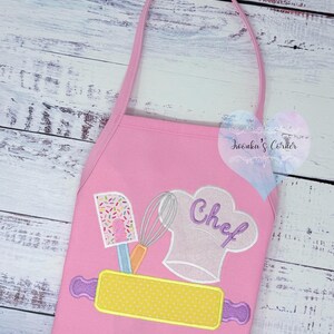 May include: Pink apron with a chef hat, rolling pin, spatula, and whisk design. The word "Chef" is embroidered on the hat. The apron has a pink strap for wearing.