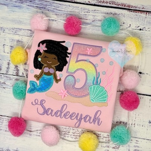 May include: Pink t-shirt with a mermaid design, the number 5, and the name "Sadeeyah" in script. The mermaid has black hair and a blue tail. A garland of colorful pom-poms surrounds the shirt.