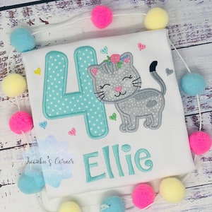 Op de afbeelding: Wit T-shirt met een geborduurd ontwerp met een grijze kat, het cijfer 4 in turquoise met witte stippen en de naam "Ellie". Het ontwerp bevat harten en bloemaccenten. Een slinger van kleurrijke pompons omringt het shirt.