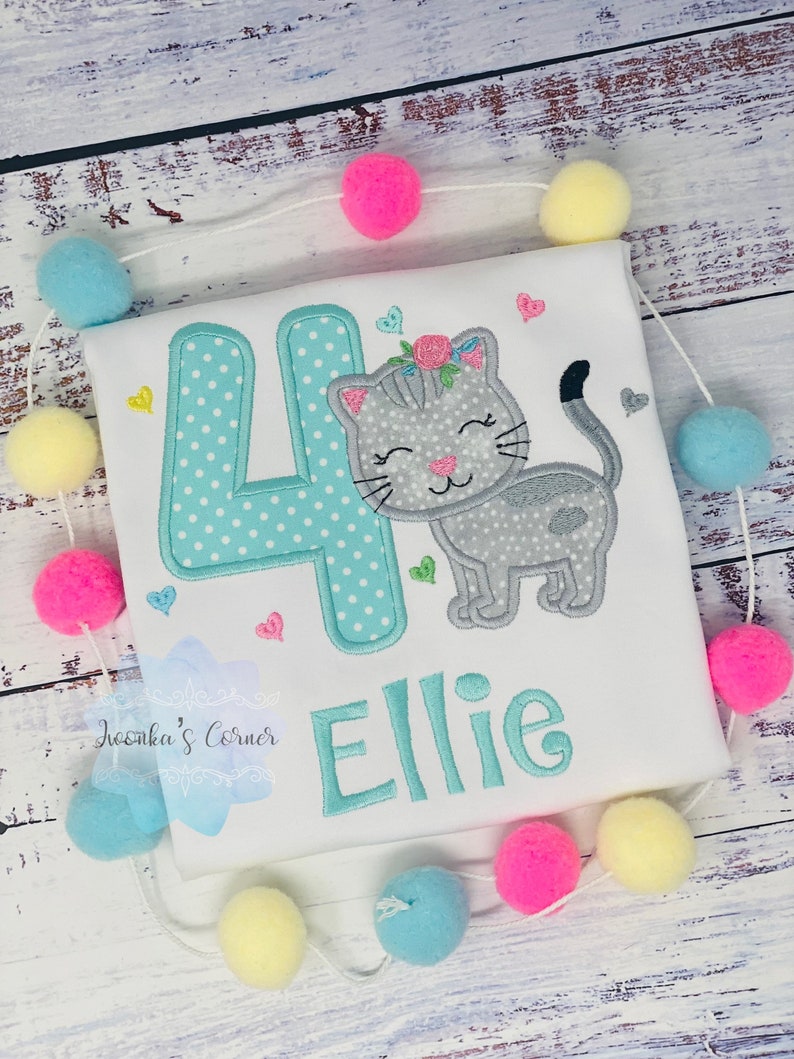 Op de afbeelding: Wit T-shirt met een geborduurd ontwerp met een grijze kat, het cijfer 4 in turquoise met witte stippen en de naam "Ellie". Het ontwerp bevat harten en bloemaccenten. Een slinger van kleurrijke pompons omringt het shirt.