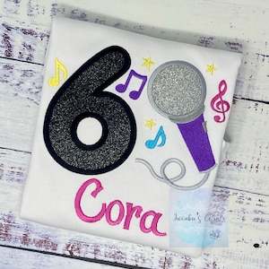 May include: A white t-shirt with a black glittery number six, a purple microphone with a silver glitter top, and musical notes. The name Cora is embroidered in pink below the design.