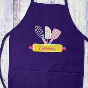 May include: Purple apron with a large pocket and adjustable straps. The apron has an embroidered design of a rolling pin with the name "Emma" on it, along with a whisk, a spatula, and sprinkles.