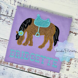Puede incluir: Una camiseta morada con un caballo marr&oacute;n con una silla de montar azul y una flor en la cabeza. El nombre "BRIDGETTE" est&aacute; bordado en turquesa en la camiseta.