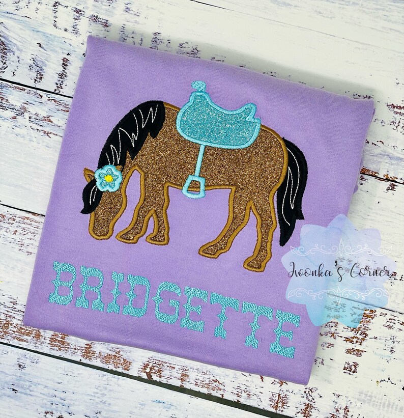 Puede incluir: Una camiseta morada con un caballo marr&oacute;n con una silla de montar azul y una flor en la cabeza. El nombre "BRIDGETTE" est&aacute; bordado en turquesa en la camiseta.