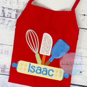 May include: Red children's apron with a yellow and white checkered rolling pin, a blue spatula, a green whisk, and a colorful ice cream scoop. The rolling pin has the name "Isaac" embroidered on it.