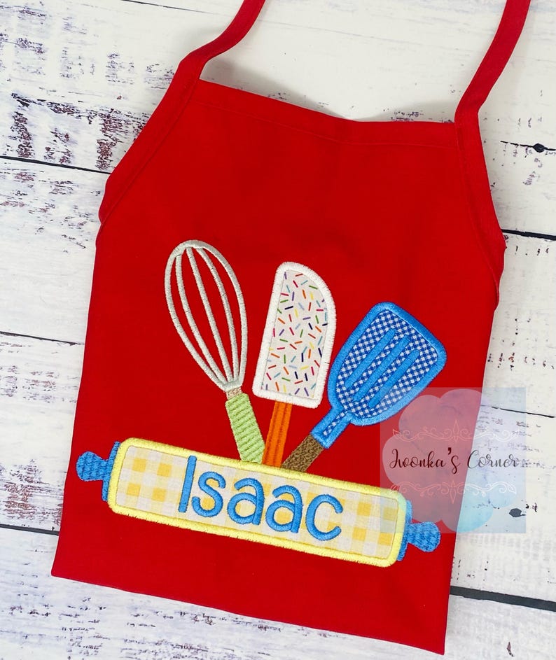 May include: Red children's apron with a yellow and white checkered rolling pin, a blue spatula, a green whisk, and a colorful ice cream scoop. The rolling pin has the name "Isaac" embroidered on it.