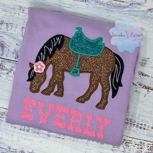 May include: Purple t-shirt with a brown horse with a teal saddle and a pink flower on its head. The text "EVERLY" is embroidered in pink on the shirt.