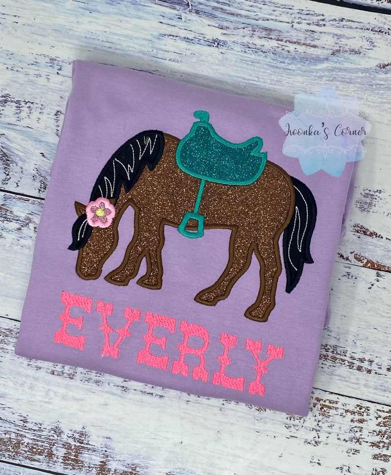 May include: Purple t-shirt with a brown horse with a teal saddle and a pink flower on its head. The text "EVERLY" is embroidered in pink on the shirt.