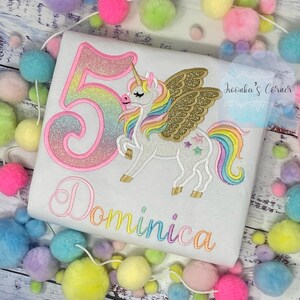 May include: White t-shirt with a colorful embroidered unicorn design, gold wings, and the number 5 in rainbow colors. The name "Dominica" is embroidered in a rainbow font. The design is surrounded by colorful pom-poms.