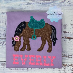 May include: Purple t-shirt with a brown horse with a teal saddle and a pink flower on its head. The name "EVERLY" is embroidered in pink below the horse.