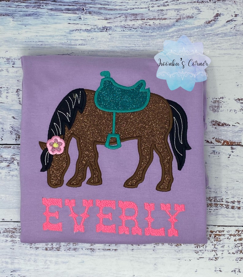 May include: Purple t-shirt with a brown horse with a teal saddle and a pink flower on its head. The name "EVERLY" is embroidered in pink below the horse.
