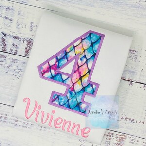 May include: White t-shirt with a purple outlined number four. The number four is filled with a mermaid scale pattern in shades of blue, pink, and yellow. The name Vivienne is embroidered in pink below the number.