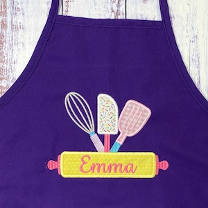 May include: Purple apron with a yellow rolling pin, a whisk, a spatula, and a frosting knife. The rolling pin has the name "Emma" embroidered on it.