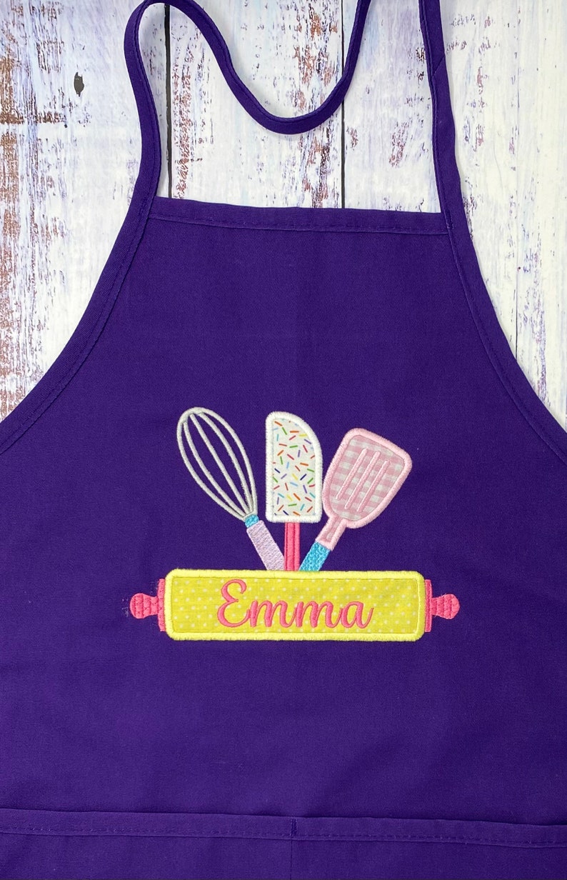 May include: Purple apron with a yellow rolling pin, a whisk, a spatula, and a frosting knife. The rolling pin has the name "Emma" embroidered on it.