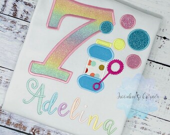 May include: A white t-shirt with a rainbow glittery number 7, a blue bottle with a pink wand, and the name "Adelina" in rainbow colors. The shirt is for a birthday celebration.