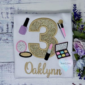 May include: White t-shirt with gold glitter number 3 and makeup-themed embroidery. Includes a nail polish bottle, makeup brushes, a compact, lipstick, and an eyeshadow palette. The name "Oaklynn" is embroidered in gold.