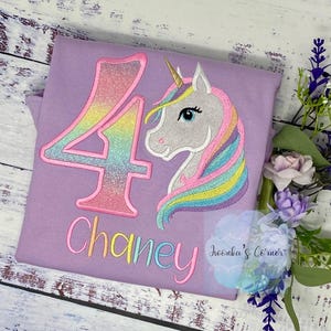 Könnte beinhalten: Lilafarbenes T-Shirt mit einer regenbogenfarbenen Glitzerzahl 4 und einem Einhorn mit einer Regenbogenmähne. Der Name "Chaney" ist unter der Zahl gestickt.