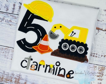 May include: A white t-shirt featuring a yellow construction vehicle, a yellow hard hat, an orange traffic cone, and the number 5 with a black road design. The text "carmine" is written below the vehicle.