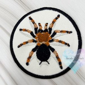 Peut inclure: Écusson brodé représentant une tarentule. L'araignée a un corps et des pattes noirs avec des marques orange et marron. L'écusson est circulaire, avec un fond blanc et une bordure noire. Pour vêtements ou accessoires.