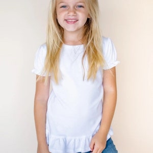 May include: A young girl wearing a white short-sleeved t-shirt with a ruffled hem and blue jeans. She has blonde hair and is smiling.