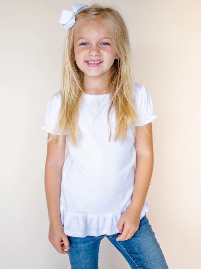 May include: A young girl wearing a white short-sleeved t-shirt with a ruffled hem and blue jeans. She has blonde hair and is smiling.