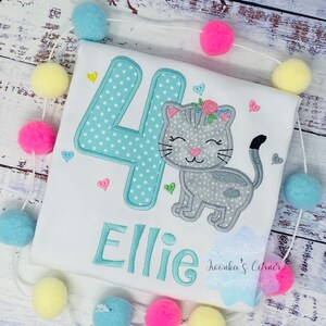 Op de afbeelding: Wit T-shirt met een turquoise nummer 4 met witte stippen en de naam "Ellie" geborduurd in turquoise. Een grijze kat met een bloemenkrans is naast het nummer geborduurd. Roze, gele en blauwe pompons zijn rond het shirt geregen.