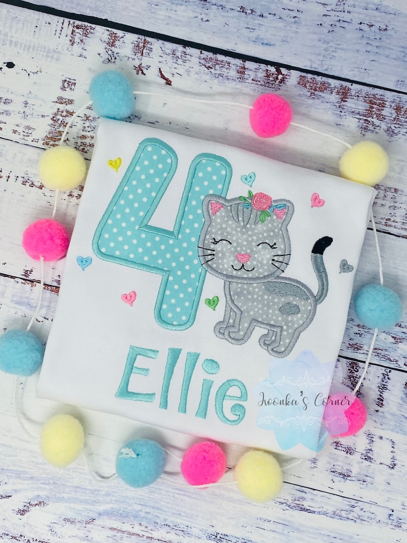 Op de afbeelding: Wit T-shirt met een turquoise nummer 4 met witte stippen en de naam "Ellie" geborduurd in turquoise. Een grijze kat met een bloemenkrans is naast het nummer geborduurd. Roze, gele en blauwe pompons zijn rond het shirt geregen.